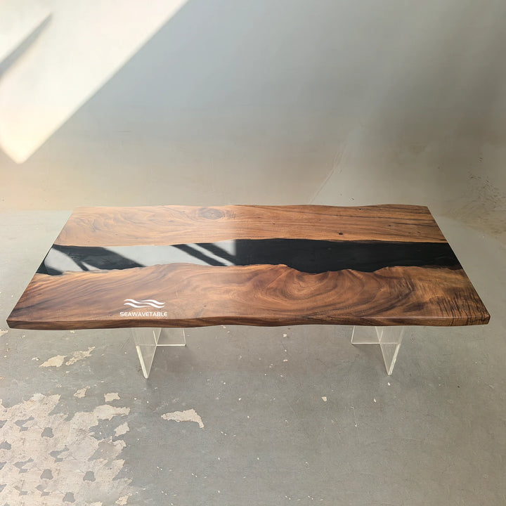 SeawaveTable--A live-edge wooden table with a glossy black resin inlay and a small white wave logo near the bottom left, supported by two clear acrylic legs in a warehouse setting.