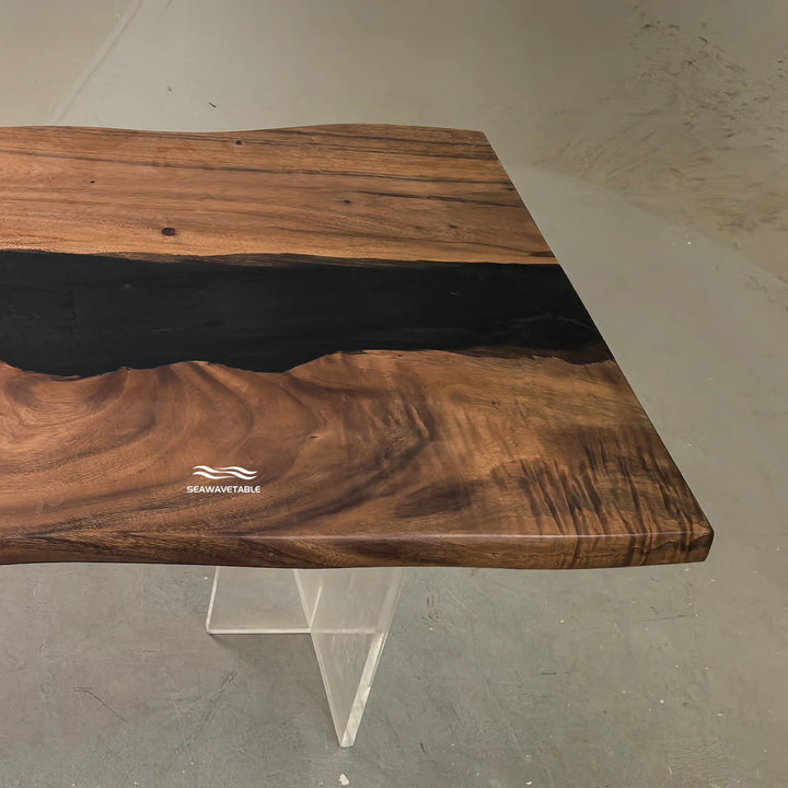 SeawaveTable-A live-edge wooden table with a rich brown wood grain and a dark black resin river running through the center, supported by a clear acrylic geometric base. The brand name 'SEAWEAVABLE' is printed on the wood.