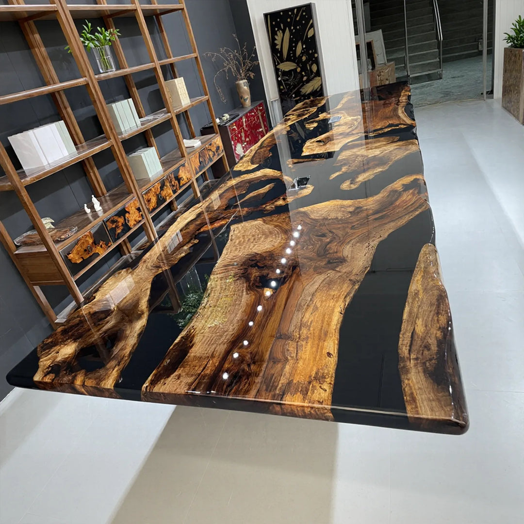 A live-edge wood conference table with a dark brown glossy surface intersected by a flowing black epoxy river, creating a striking contrast between organic grain and modern resin, surrounded by beige upholstered chairs in a contemporary meeting space with panoramic mountain views.