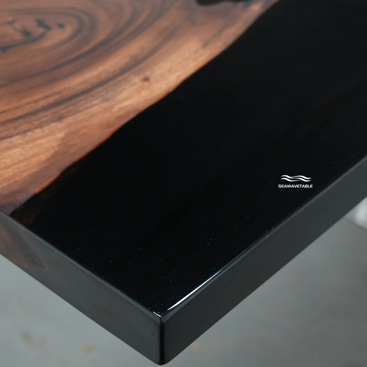 Close-up of a table corner showing a glossy black edge with a natural brown wood grain top, branded with the SAWANTABLE logo.