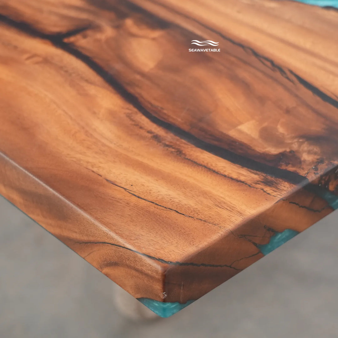 Close-up of a live-edge wooden tabletop with rich warm brown grain and small turquoise resin inclusions at the corners, photographed on a neutral concrete floor. The brand name SeaWhitetable is printed near the top edge.