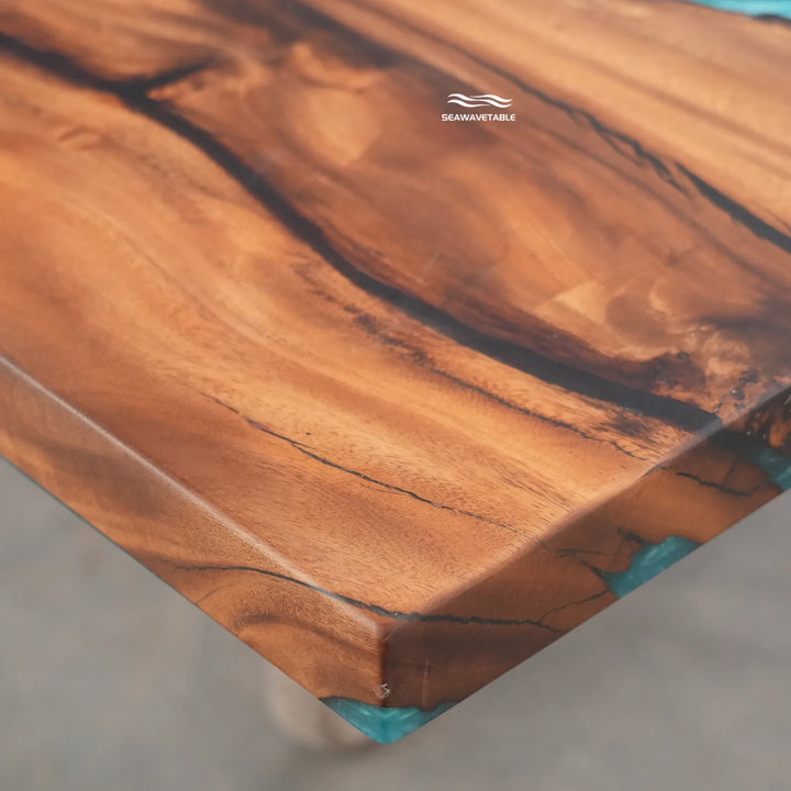 Close-up of a live-edge wooden tabletop with rich warm brown grain and small turquoise resin inclusions at the corners, photographed on a neutral concrete floor. The brand name SeaWhitetable is printed near the top edge.