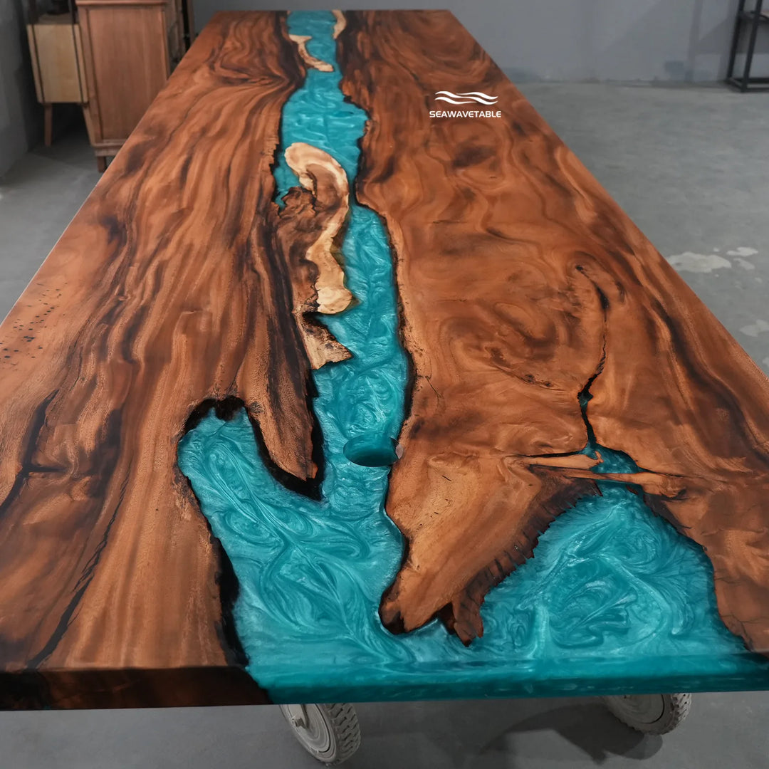A large live-edge wooden table with a turquoise river design running down the center, photographed in a workshop setting. The wood has rich brown and black grain patterns, and the turquoise resin inlay is highly detailed and swirling.