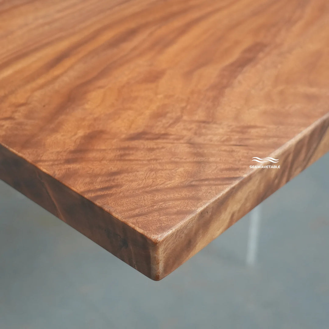 Close-up of a wooden tabletop corner showing rich grain and a warm medium-brown finish, with a small visible SHAWMATABLE watermark and a hint of a light-colored table leg below.