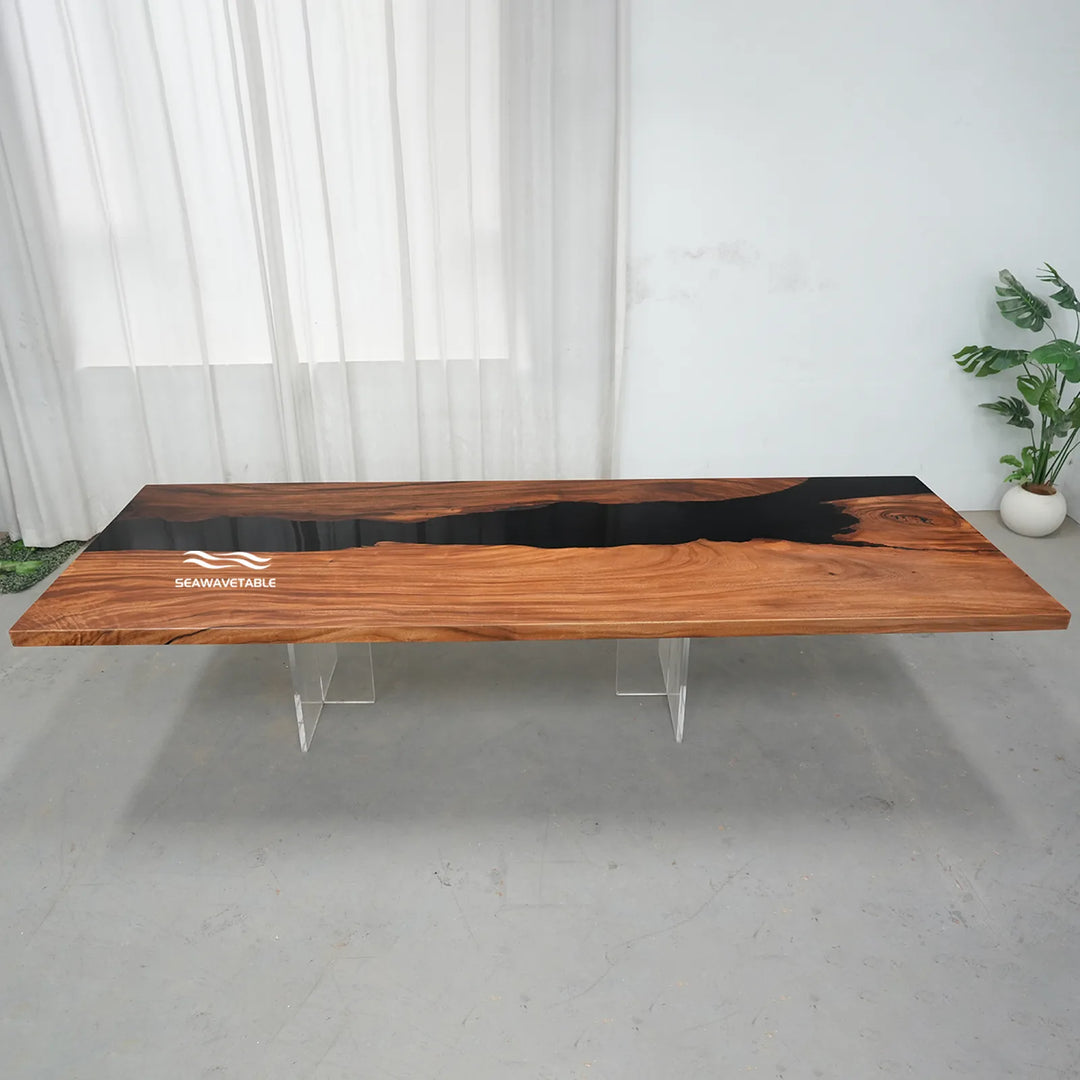 A live-edge wooden table with a glossy black resin river inlay, supported by clear acrylic legs, set in a minimalist room with white curtains and a potted plant.