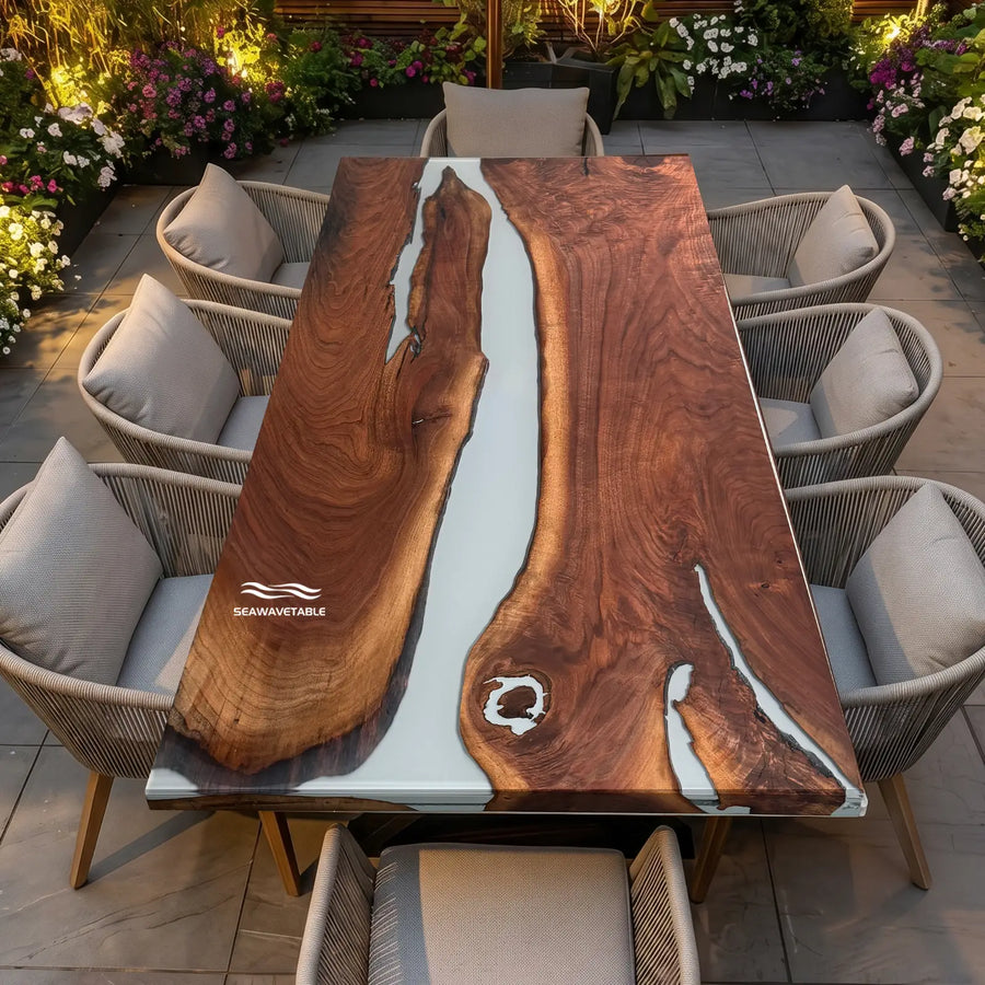 A large rectangular live-edge black walnut epoxy dining table with a white resin river channel, surrounded by gray woven patio chairs on a tiled patio with lush green plants and natural sunlight - SeawaveTable
