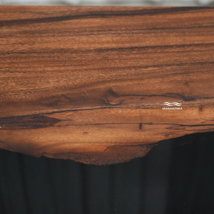 Close-up of a live-edge wooden tabletop with rich warm brown grain and a small visible SEAWAVE TABLE logo, set against a dark background.