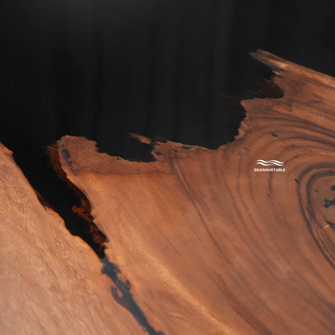 Close-up of a live-edge wooden tabletop with rich warm brown wood grain and dark resin channels, photographed against a dark background. A small 'SEAANTICABLE' logo appears on the wood surface.