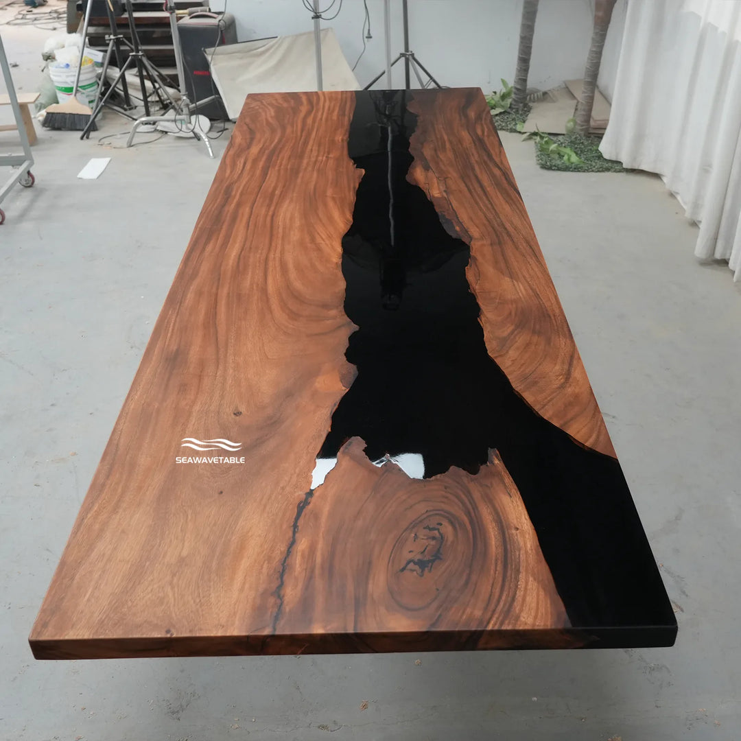 A large live-edge wooden table with a rich brown wood grain and a glossy black resin channel running down the center, photographed in a workshop setting.