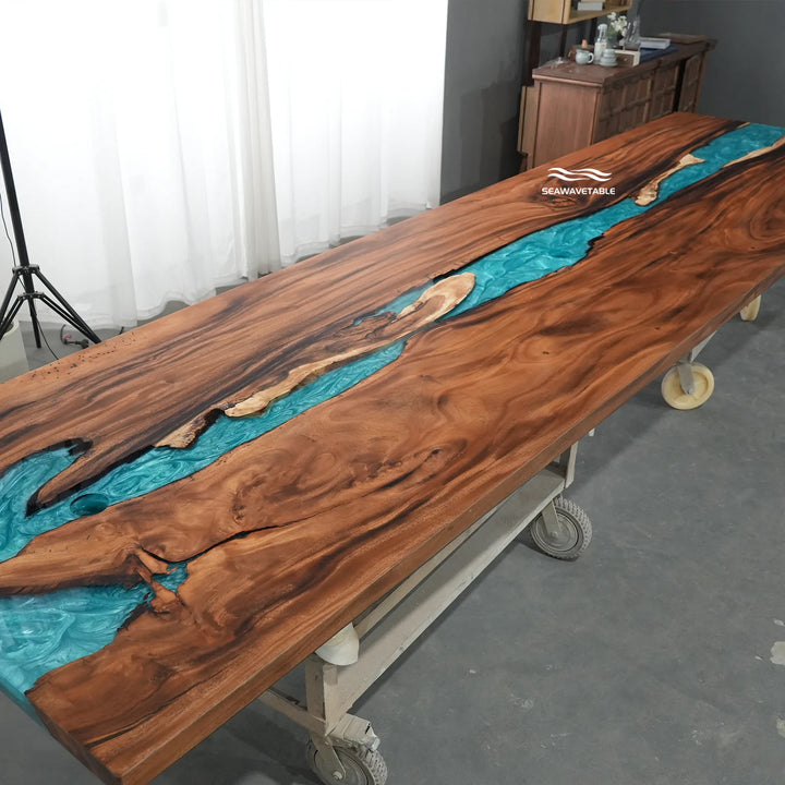 A large live-edge wooden table on a wheeled metal frame, featuring a rich brown wood grain with a vibrant turquoise resin channel running through the center. The table is photographed in an indoor studio or workshop setting with curtains and a gray concrete floor.