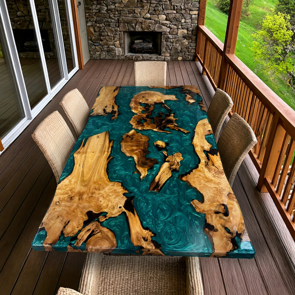 Seawavetable-A rectangular live-edge wooden table with a glossy teal resin finish and natural wood inclusions, shown on a wooden deck with woven dining chairs. A stone fireplace and railing are visible in the background.