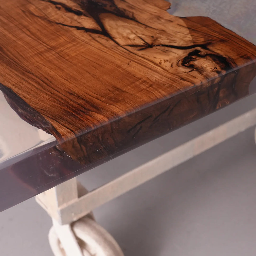 Close-up of a live-edge wooden tabletop slab with rich brown wood grain and dark natural fissures, mounted on a clear glass board over a white rolling stand.